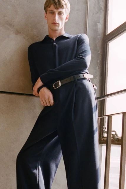 A blond man wearing a navy polo shirt and navy trousers, leaning against a wall with a window with metal bars, indoor setting.