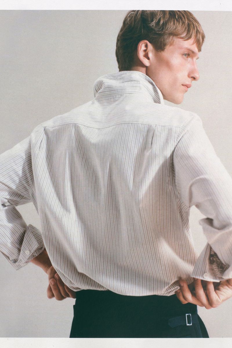 Fashionable young adult man poses in a collared pinstripe shirt and dark pants against a light neutral backdrop in a studio setting.