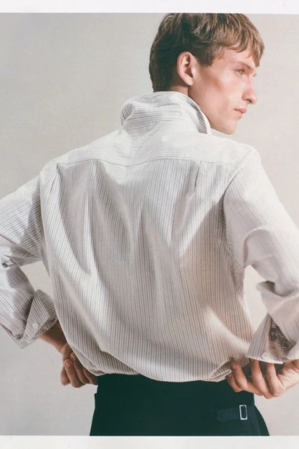 Fashionable young adult man poses in a collared pinstripe shirt and dark pants against a light neutral backdrop in a studio setting.