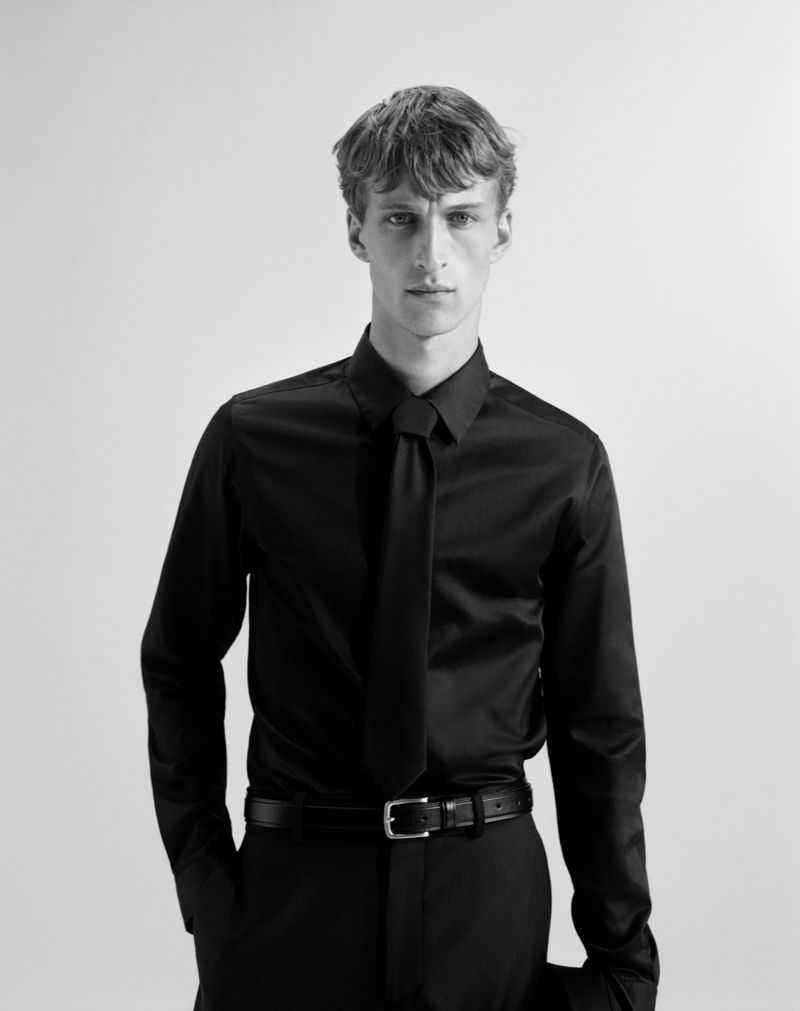 Monochrome portrait of a light-skinned adult man in a black dress shirt and tie, standing indoors against a neutral background.