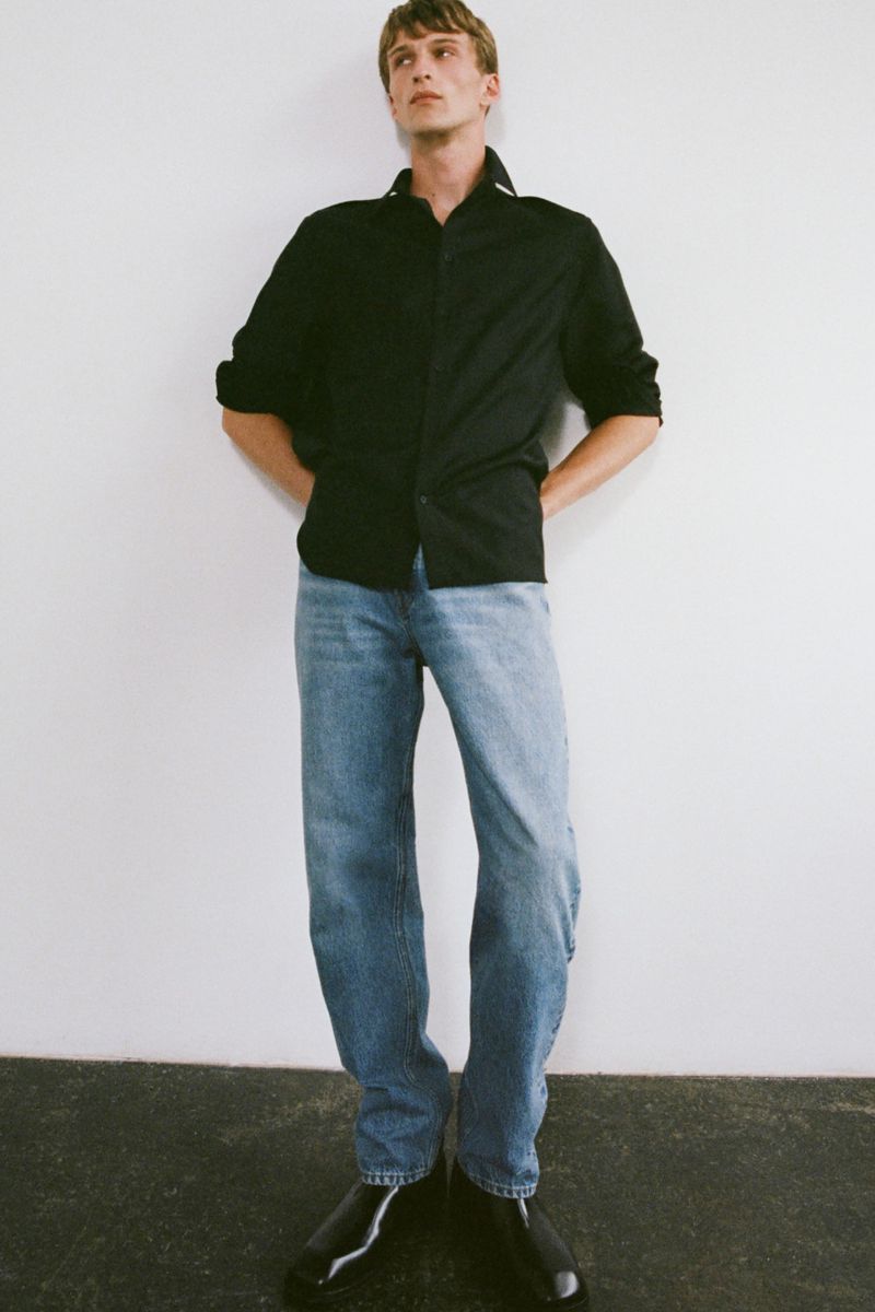 Full-length shot of a young adult man in a black button-down shirt, blue jeans, and black shoes standing against a white wall.