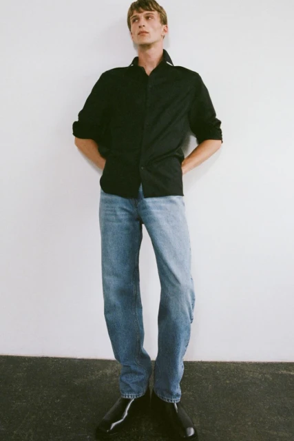 Full-length shot of a young adult man in a black button-down shirt, blue jeans, and black shoes standing against a white wall.