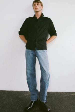 Full-length shot of a young adult man in a black button-down shirt, blue jeans, and black shoes standing against a white wall.