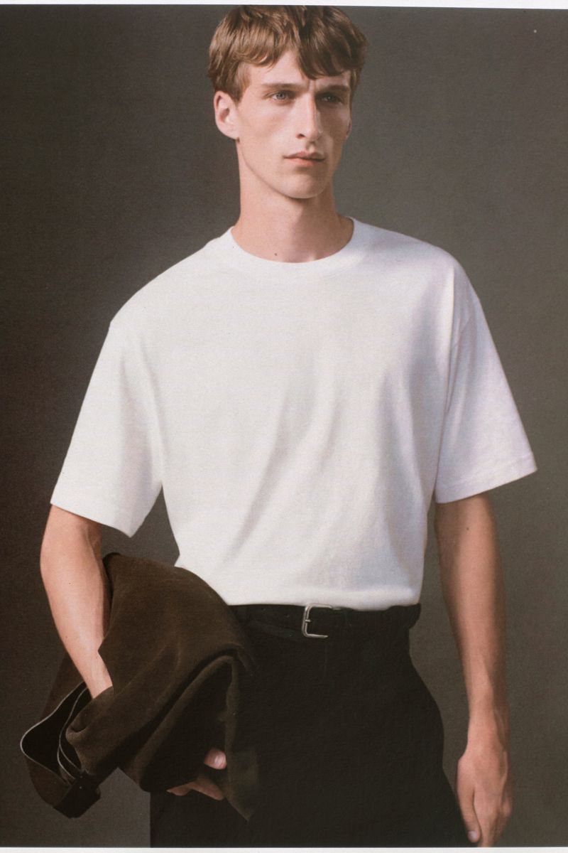 Model Vasko Luyckx wearing a white crewneck t-shirt and black trousers against a neutral grey studio backdrop for COS.