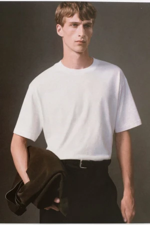Model Vasko Luyckx wearing a white crewneck t-shirt and black trousers against a neutral grey studio backdrop for COS.