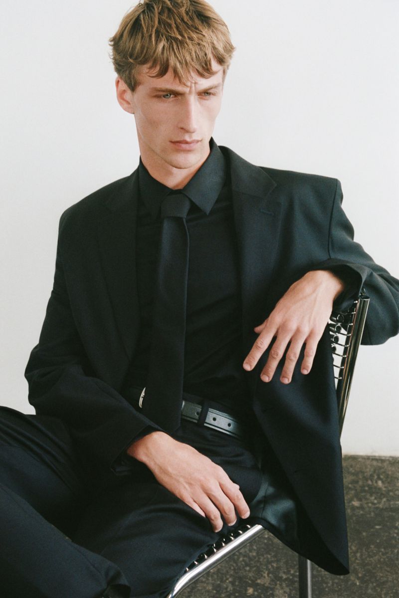 Portrait of a male model in a black suit and tie, sitting in a chair against a plain white background in a professional studio.