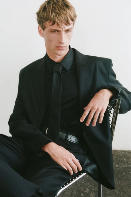 Portrait of a male model in a black suit and tie, sitting in a chair against a plain white background in a professional studio.