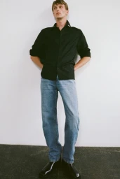 Model Vasko Luyckx leans against a white wall wearing a black button-down shirt, light-wash blue jeans, and black leather shoes.