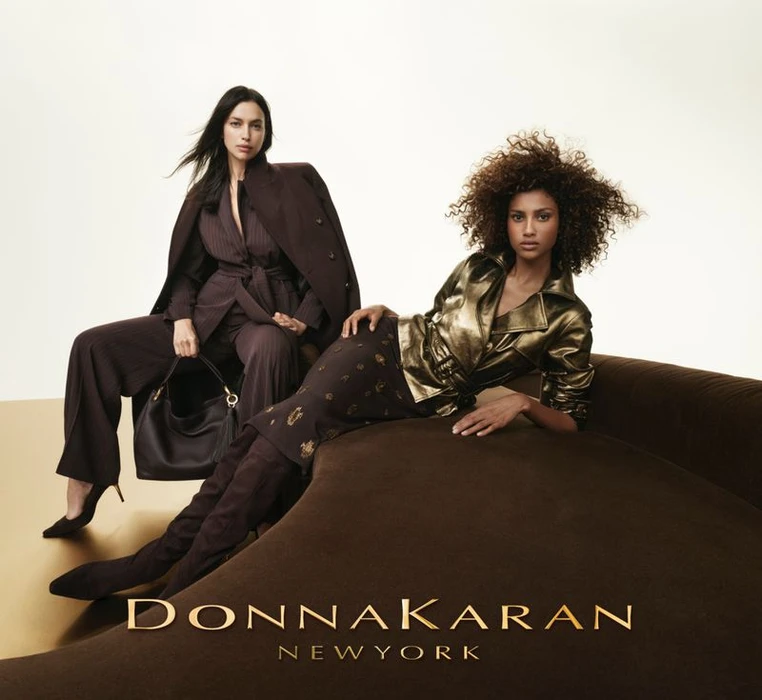 Two female models posing for Donna Karan, one seated in a brown suit and one reclining on a velvet sofa against a clean studio backdrop