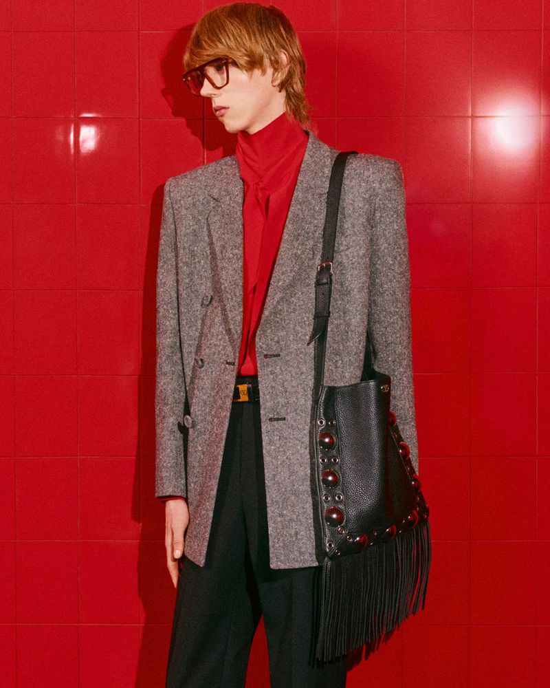 Adult male fashion model in a grey blazer, red turtleneck, and black slacks, with a handbag, posing against a red tile background.