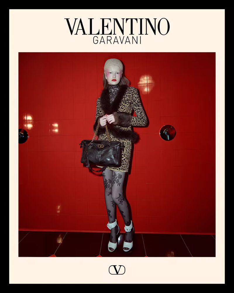 Valentino Garavani fashion: Woman in animal-print dress, fur-trimmed jacket, patterned tights, platform heels, and black purse, red backdrop.