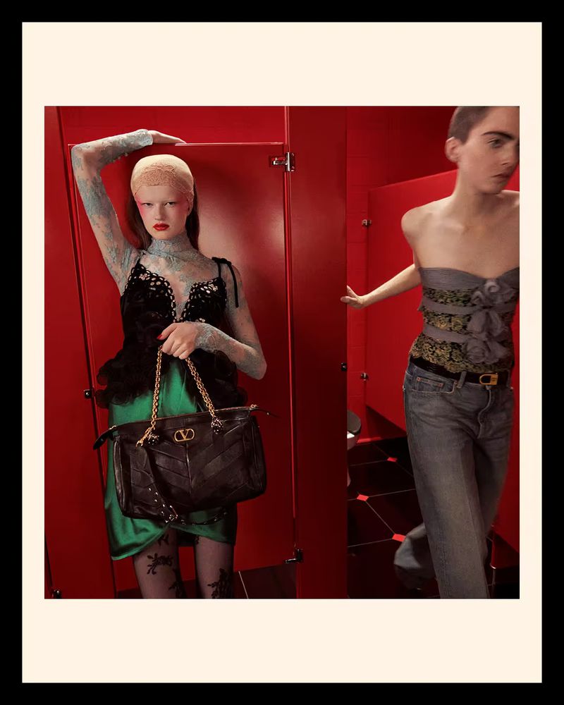 Two women in a red-walled interior setting. One woman holds a black purse with gold chain, while the other stands between red doors.