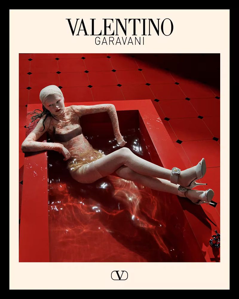 Fashion shot of a woman in a bathtub filled with red liquid, wearing a light-colored dress and high heels. Valentino Garavani logo visible.