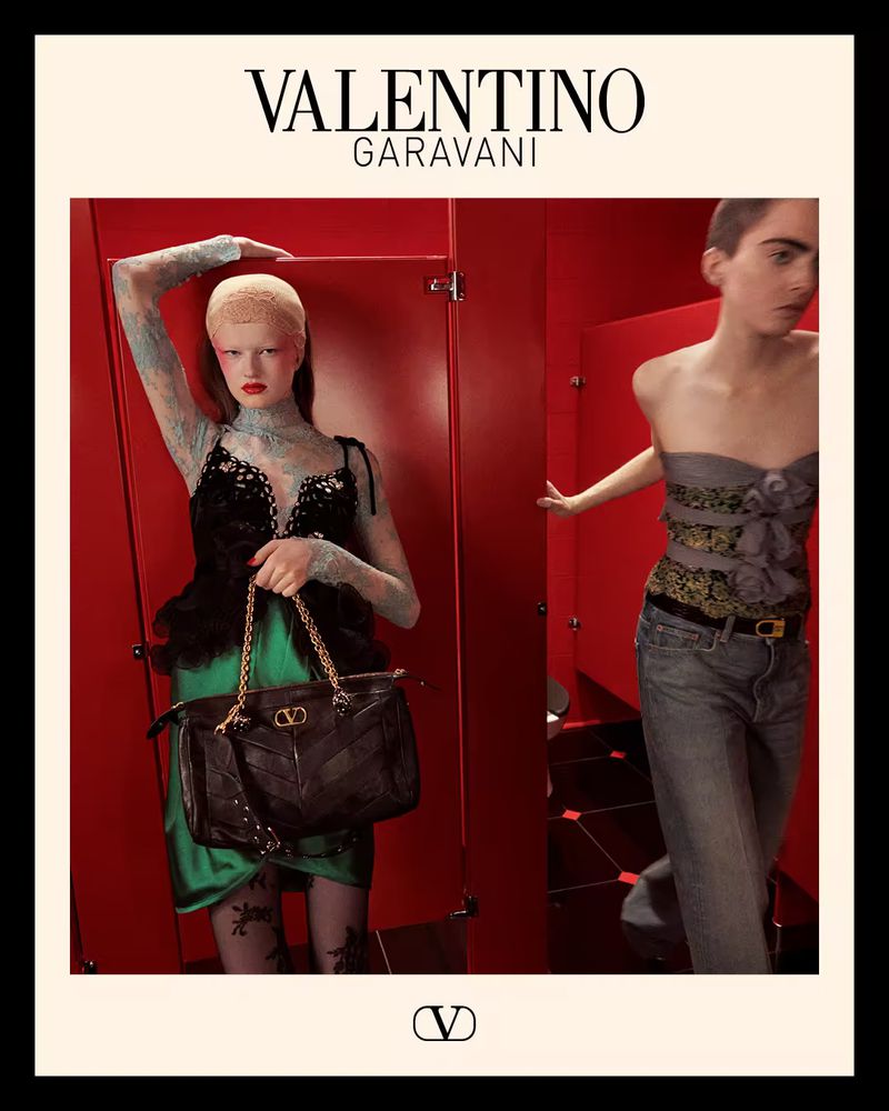 Two fashion models pose against a red wall in a studio. One carries a Valentino Garavani handbag and wears a green skirt.