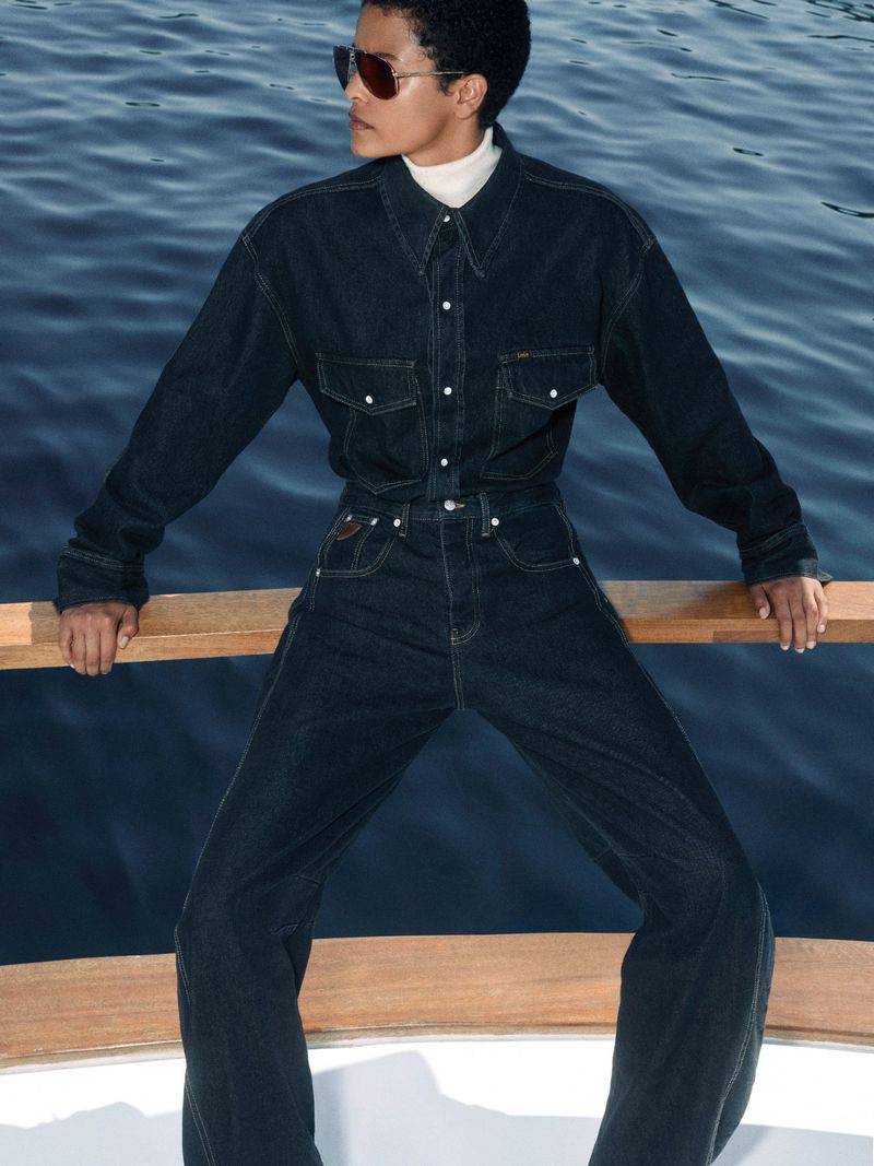Adult posing on a boat wearing sunglasses and a denim outfit, captured in a medium shot on a bright day with blue water.
