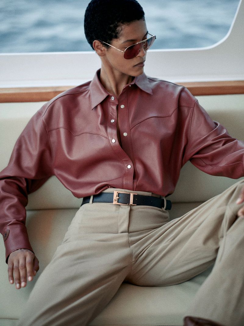 Woman in aviator sunglasses and a maroon leather shirt sitting on a boat in a fashion shot, complemented by cream-colored slacks and a black belt.
