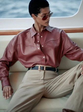 Woman in aviator sunglasses and a maroon leather shirt sitting on a boat in a fashion shot, complemented by cream-colored slacks and a black belt.