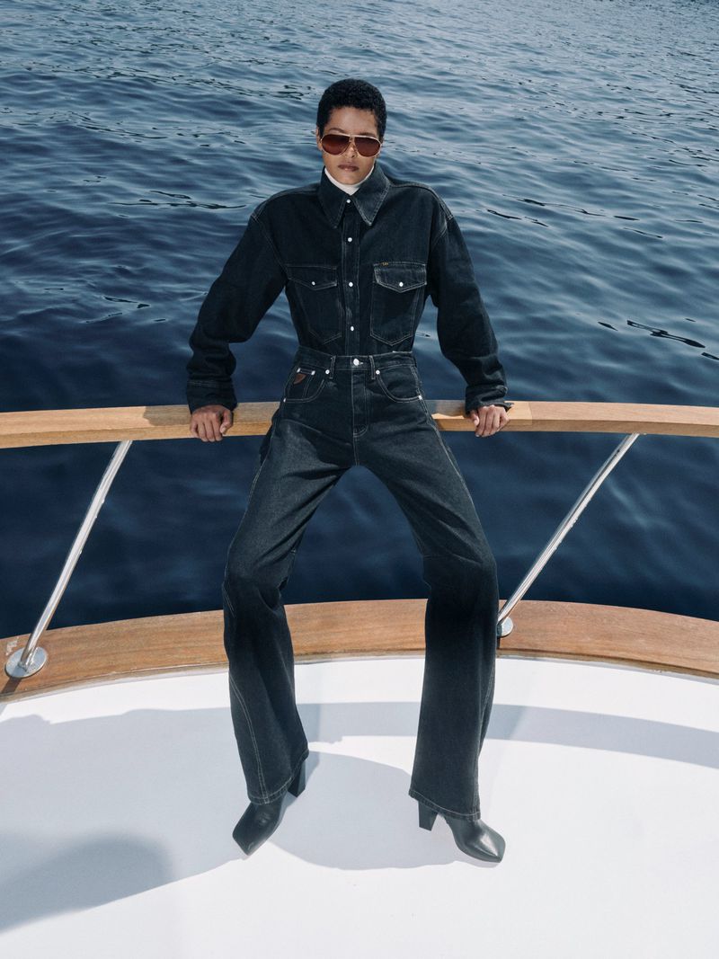 A person in a dark denim suit and tinted aviator sunglasses is sitting on the railing of a boat on a bright, sunny day at sea.