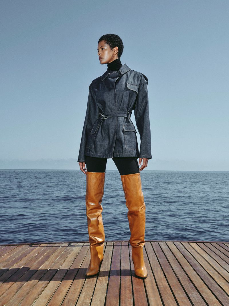 A woman models a denim jacket, black turtleneck, and thigh-high boots on a wood deck overlooking the sea against a blue sky.