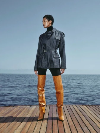 A woman models a denim jacket, black turtleneck, and thigh-high boots on a wood deck overlooking the sea against a blue sky.