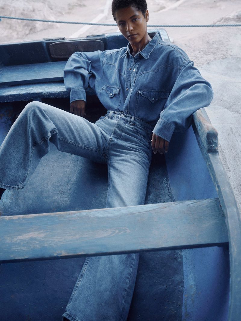 Adult female fashion model poses in a denim shirt and wide-leg jeans inside of a bright blue rowboat, resting on light tan sand.