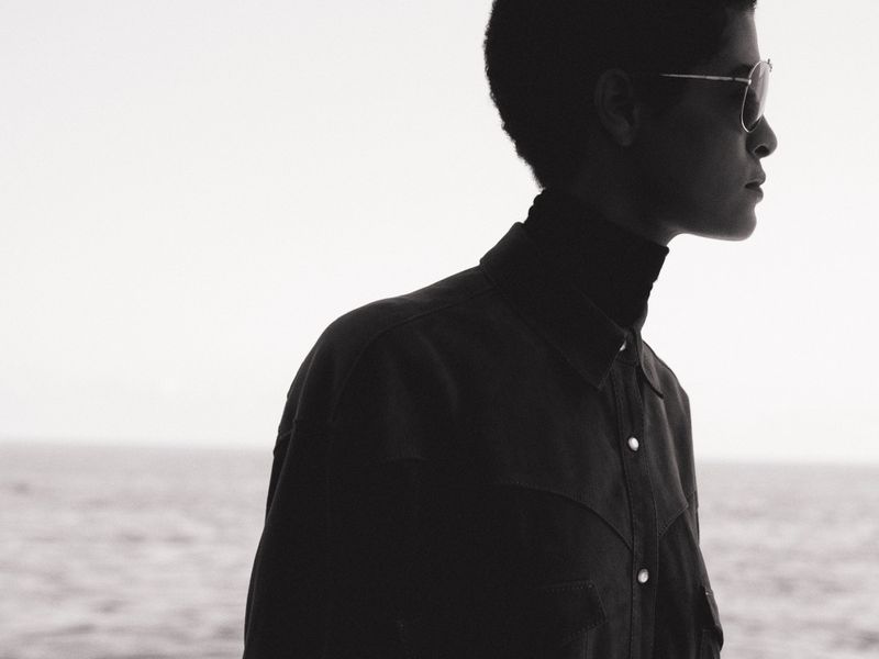 Adult male wearing sunglasses and a dark jacket stands in profile against a neutral sky and ocean backdrop, showcasing fashion and style.