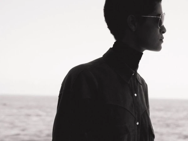 Adult male wearing sunglasses and a dark jacket stands in profile against a neutral sky and ocean backdrop, showcasing fashion and style.