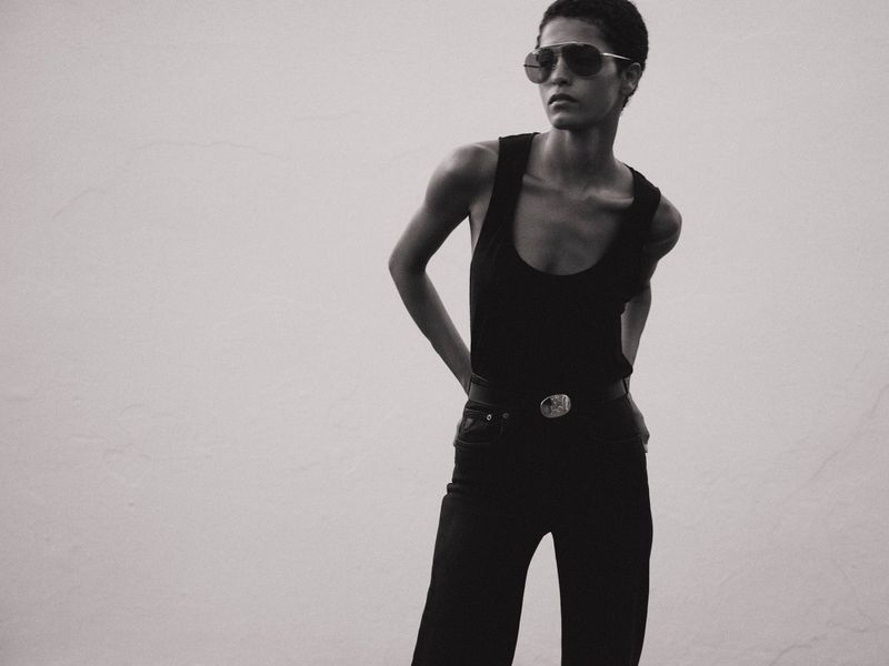 Monochrome studio portrait of a stylish adult female wearing sunglasses, black tank top, jeans and belt with a buckle against white backdrop.