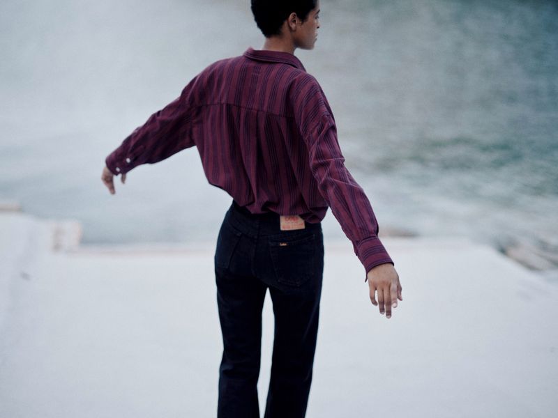 Woman in a striped shirt and black jeans stands with arms outstretched against a muted turquoise water backdrop, back to camera.