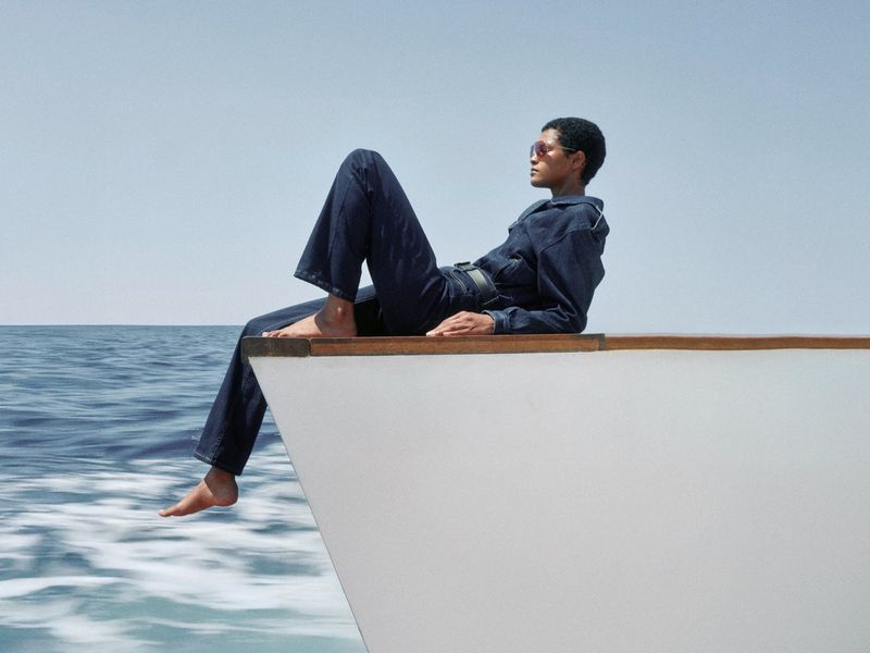 An adult is relaxing on a white boat in stylish blue denim jumpsuit, featuring the blue sea, clear sky, and boat wake.
