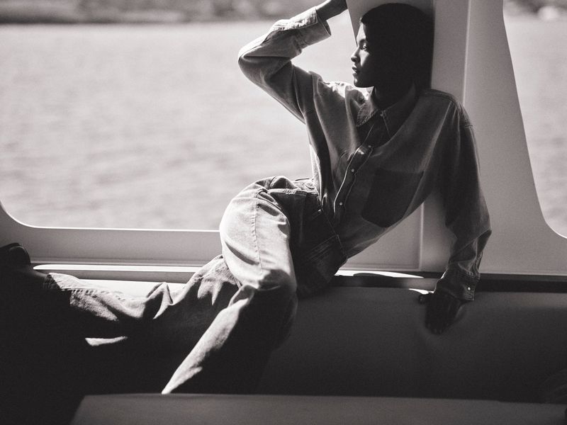 Laiza de Moura reclines in a denim shirt and jeans next to a sunlit boat window in black and white.