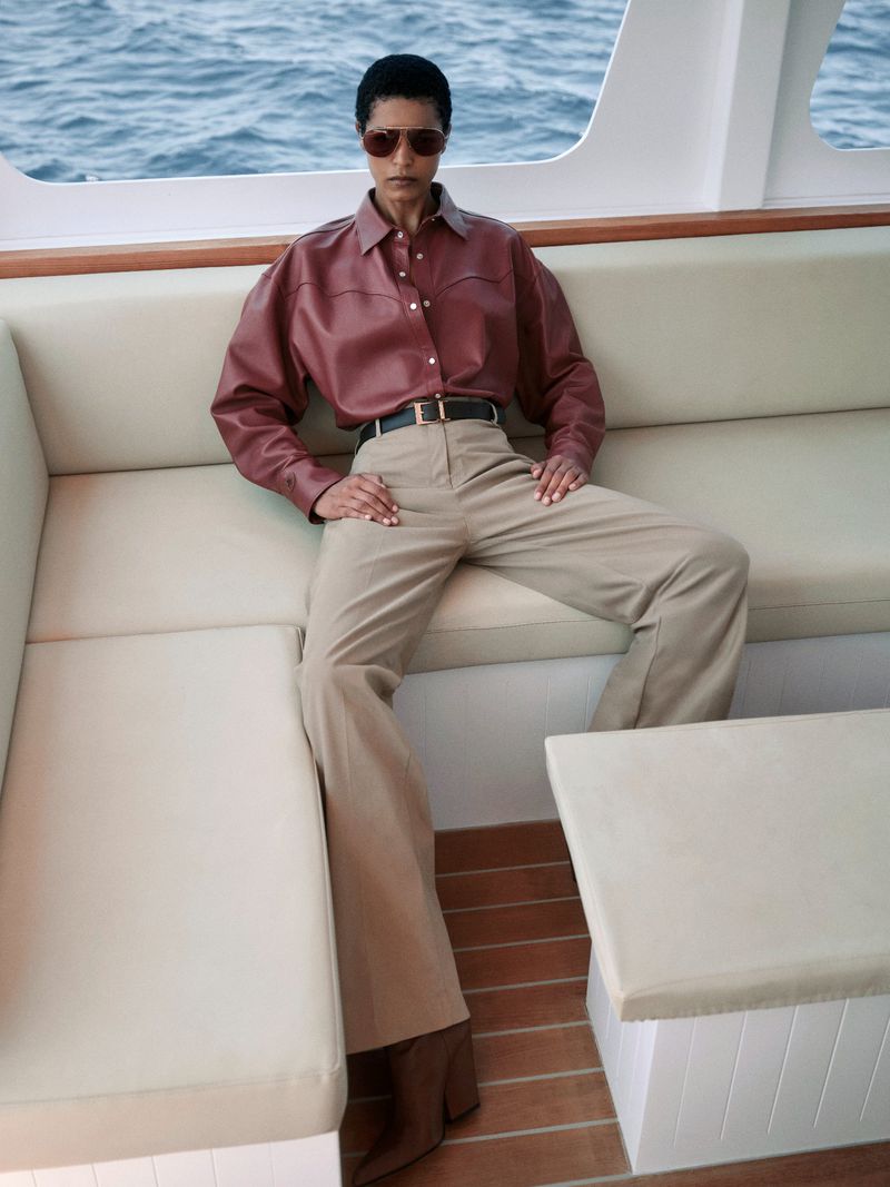 A woman in a maroon shirt and khaki pants poses on a white cushioned boat with the ocean visible in the background on a sunny day.