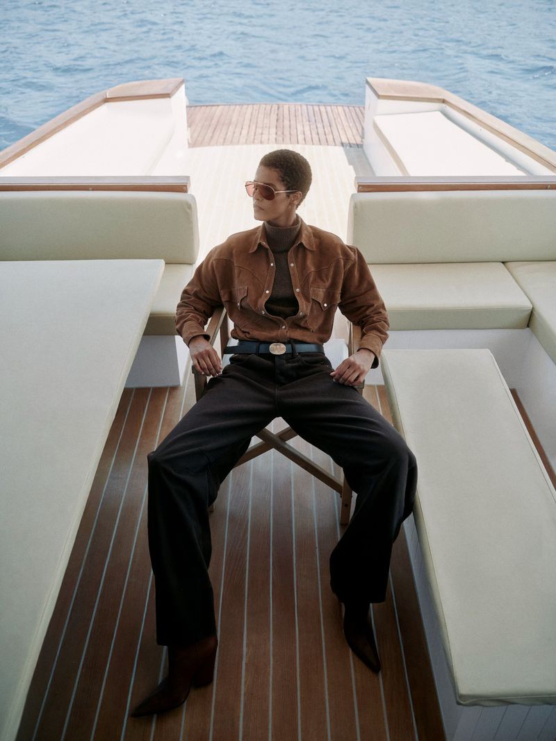 A woman wearing sunglasses and a brown suede jacket poses while sitting on a chair on the back of a boat on open water.