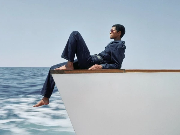 Model Laiza de Moura wearing a dark denim jumpsuit seated barefoot on the bow of a moving boat against an open ocean horizon.