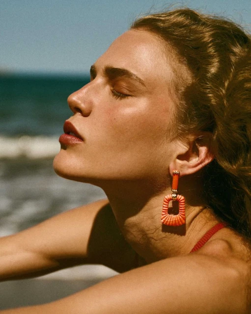 Close up portrait of an adult female wearing orange square earrings on a beach with sunlight and ocean waves in the background.