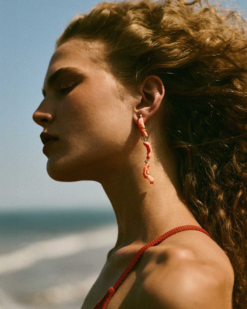 Portrait of a woman with curly hair wearing coral earrings, posing on a beach with waves, against a blue sky on a sunny day.