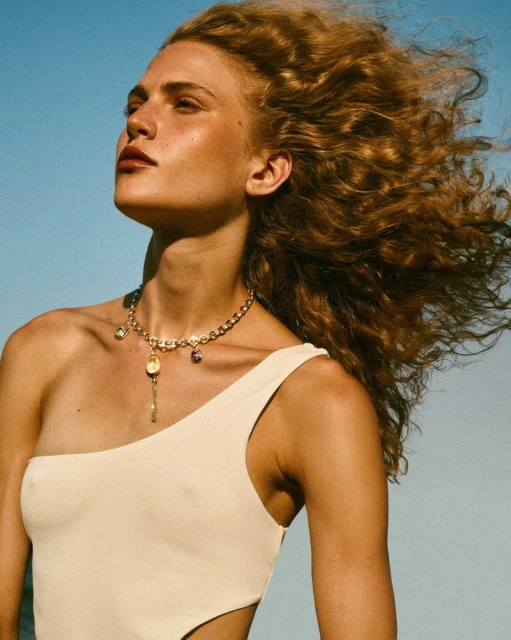 Woman with wind-blown curly hair wears asymmetric one-piece bathing suit and gold charm necklace against light blue sky in a fashion shot.