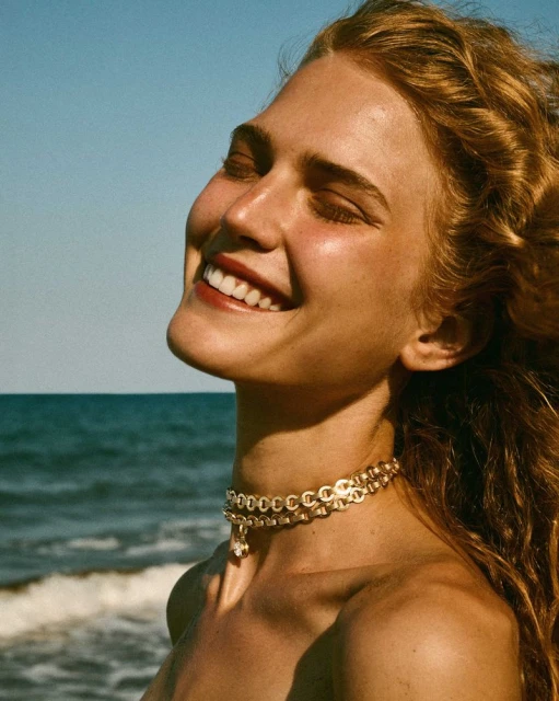 Close-up of a woman with auburn hair, smiling against a backdrop of a blue sky and ocean, wearing a gold choker necklace.