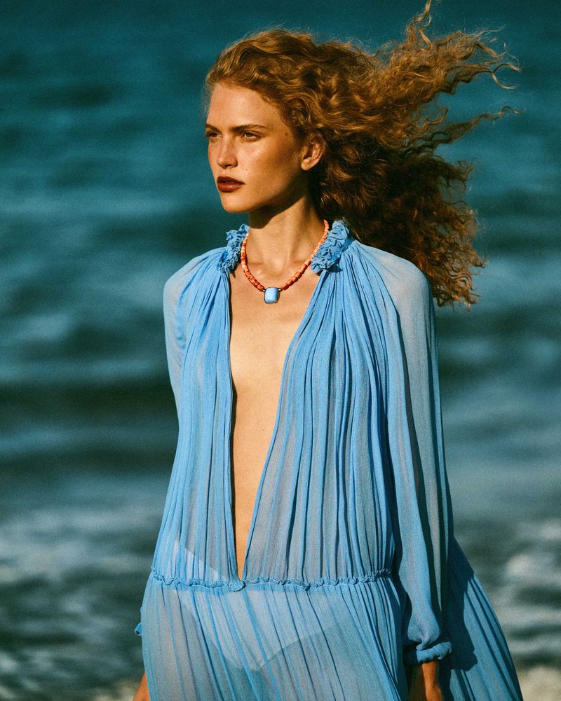 Woman with windblown curly auburn hair wearing a sheer pleated sky-blue dress and a beaded necklace against an ocean backdrop.