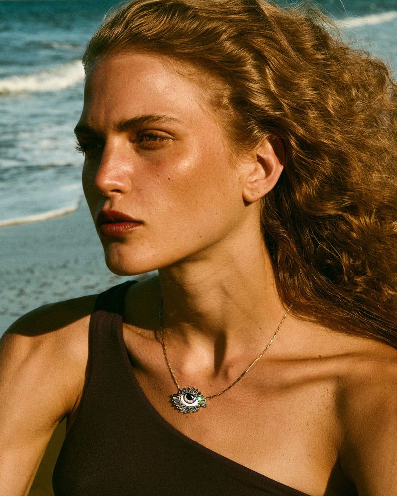 Close-up of a light-skinned woman with wavy blonde hair wearing a brown one-shoulder top and an eye-shaped pendant necklace at the beach.