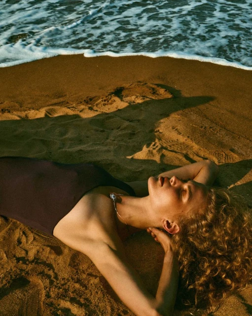 Woman in swimwear laying on a sandy beach with wavy ocean water in the background; golden hour lighting