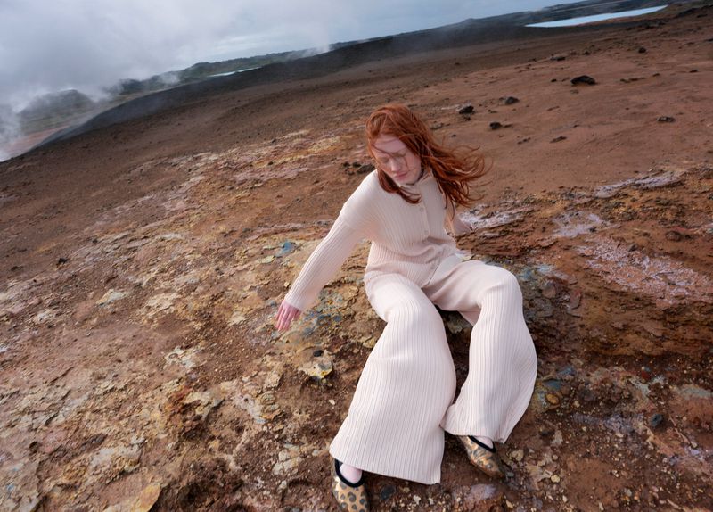 Redhead woman in matching cream knit sweater and pants posing on rocky terrain with distant steaming geysers and cloudy skies.