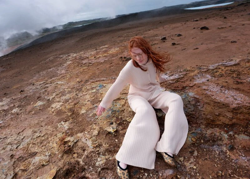 Redhead woman in matching cream knit sweater and pants posing on rocky terrain with distant steaming geysers and cloudy skies.