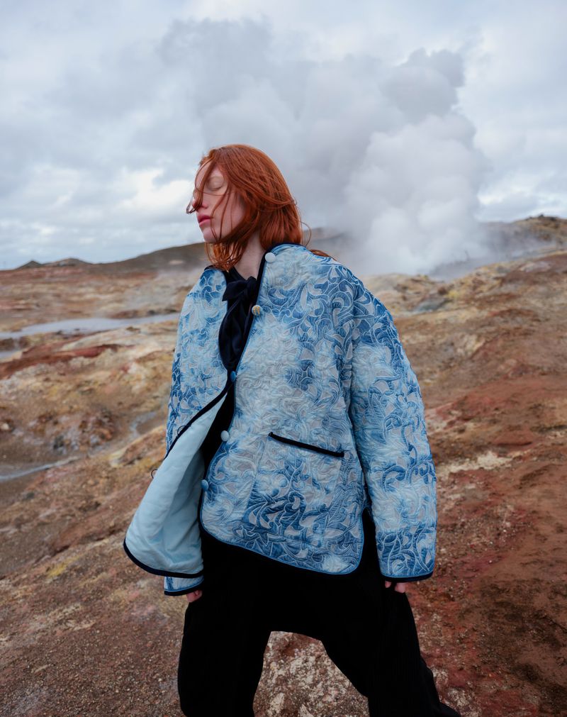 Adult woman with closed eyes stands in a landscape wearing a floral pattern jacket against a background of steam vents and clouds.