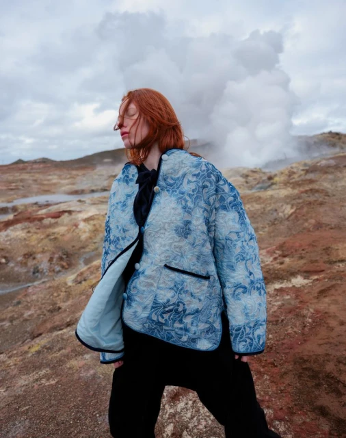 Adult woman with closed eyes stands in a landscape wearing a floral pattern jacket against a background of steam vents and clouds.