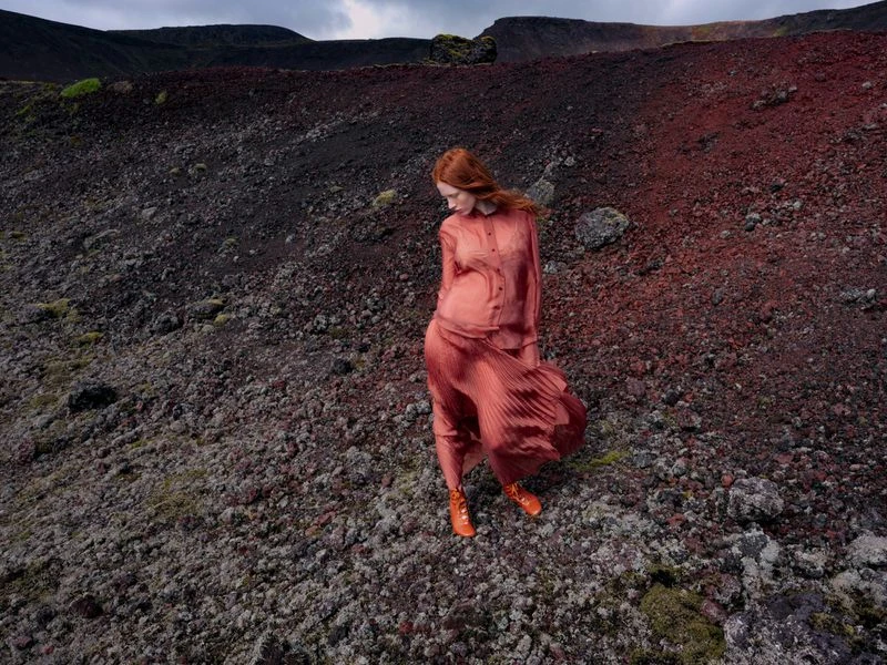 Adult woman with red hair stands in windswept red dress in a rocky, volcanic landscape under an overcast sky.