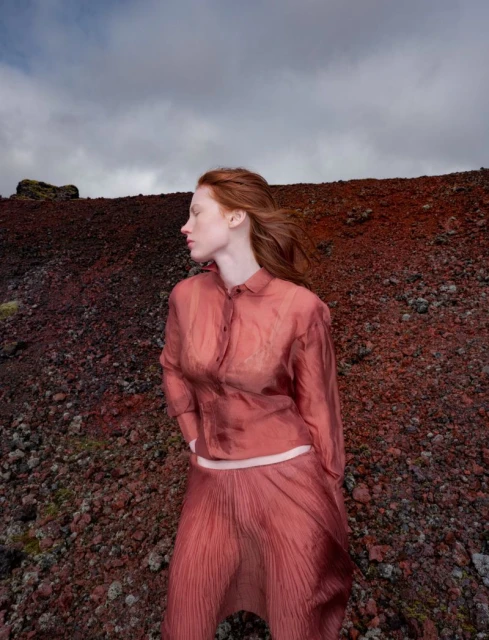 Adult woman with red hair stands among red rocks in a red blouse and skirt. Cloudy sky. Fashion editorial, nature background.