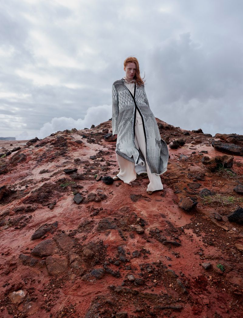 Red-haired woman in flowing grey patterned coat and white pants poses on a red rocky landscape beneath a cloudy sky for a fashion editorial.