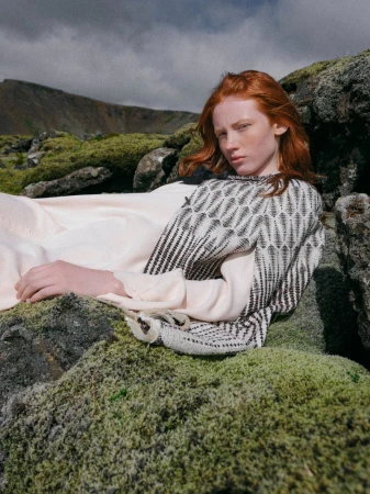 Redhead model in patterned sweater lays on mossy rocks with a mountain in the background in an outdoor fashion editorial shot.