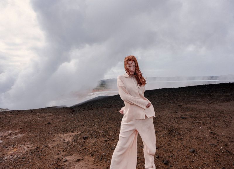Fashion photography showcasing a young woman with red hair in a beige outfit, posing against a volcanic landscape with steam in background.